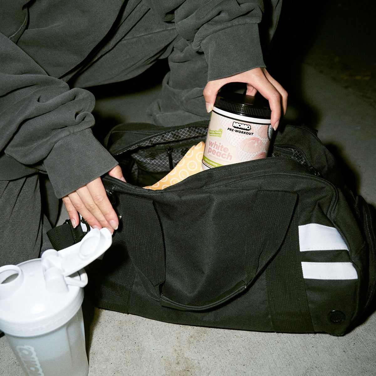 gym girl pulling out a tub of MOMO white peach pre-workout from her black duffle bag early in the morning