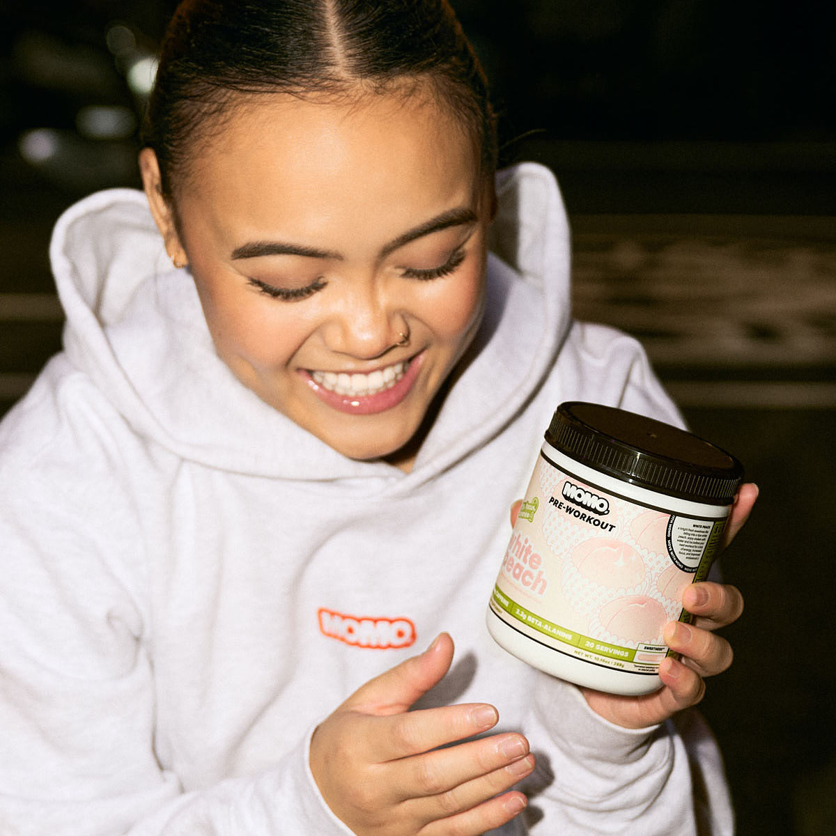 gym girl smiling in the gym, holding a tub of MOMO white peach pre-workout