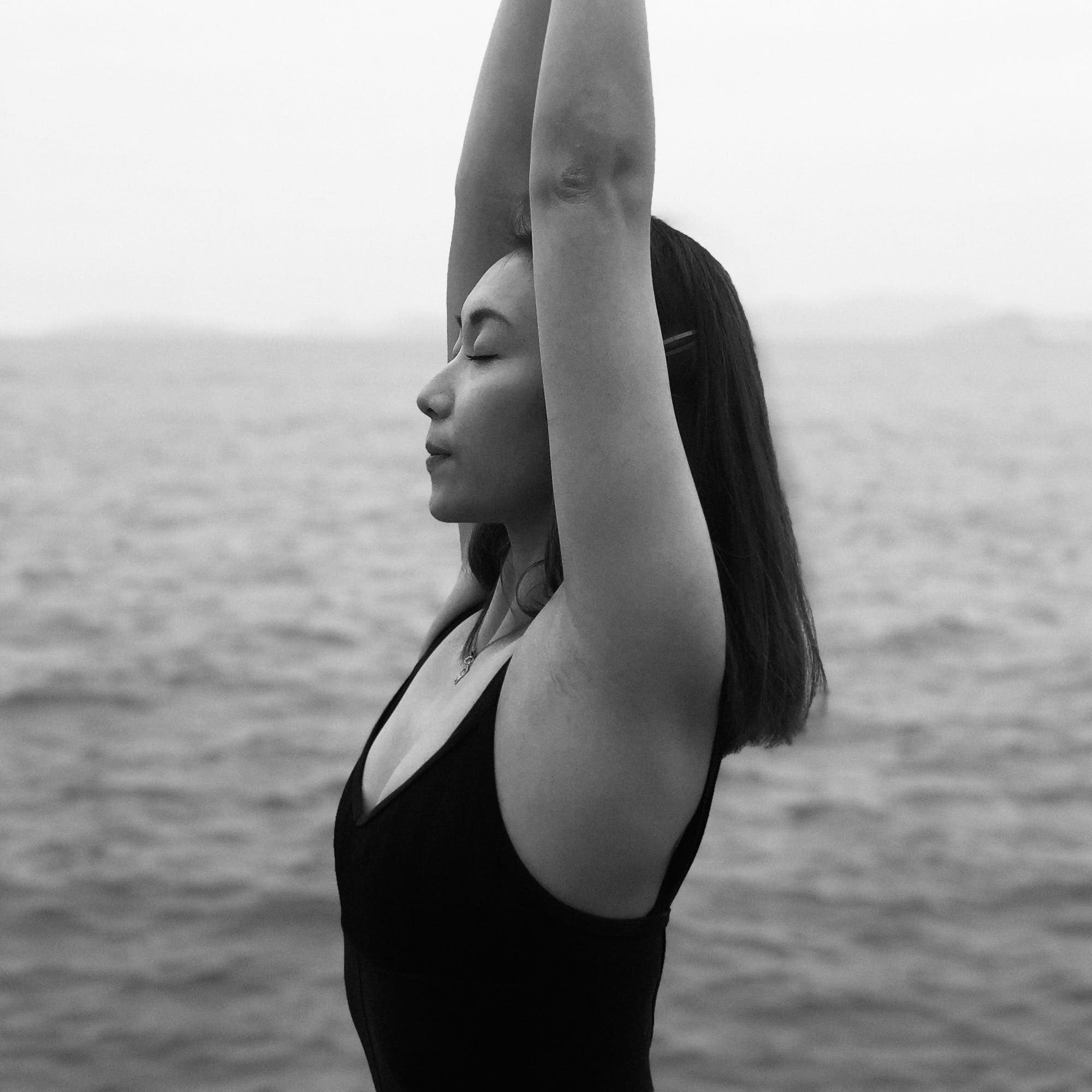 girl with her eyes closed by the beach, breathing deeply with her arms above her head