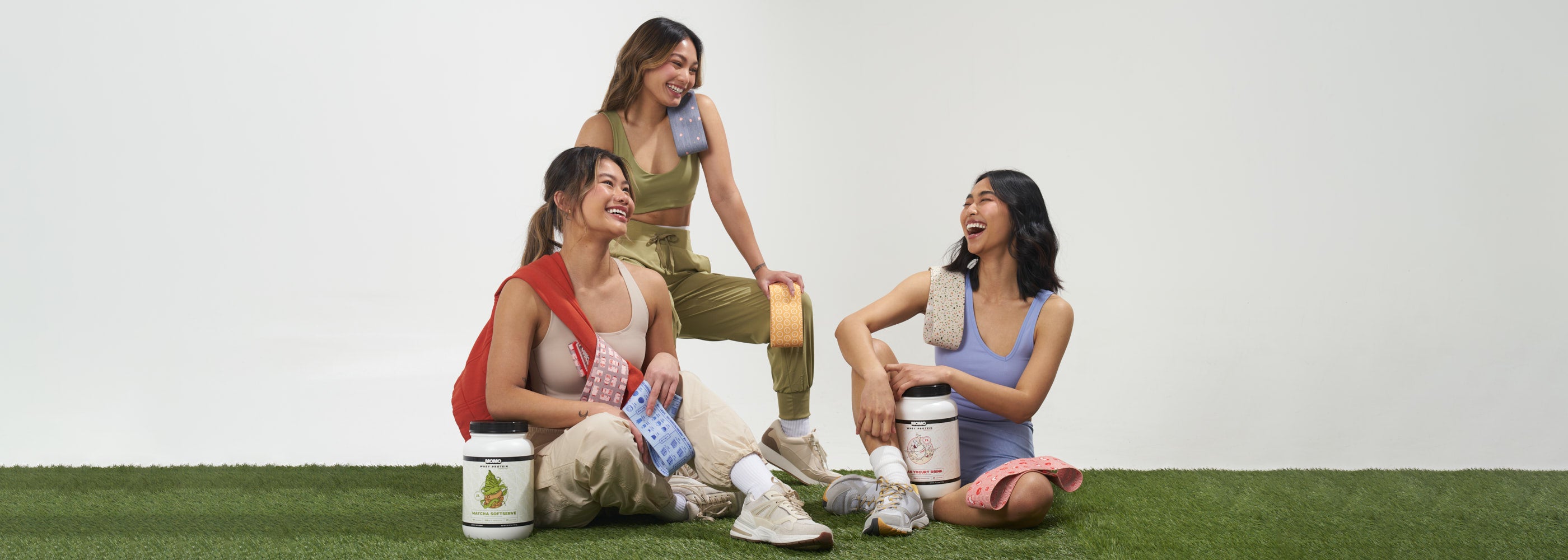 3 girls sitting and laughing with cute asian inspired fabric resistance bands, a matcha softserve whey protein, and an Asian Yogurt Whey Protein. All MOMO branded