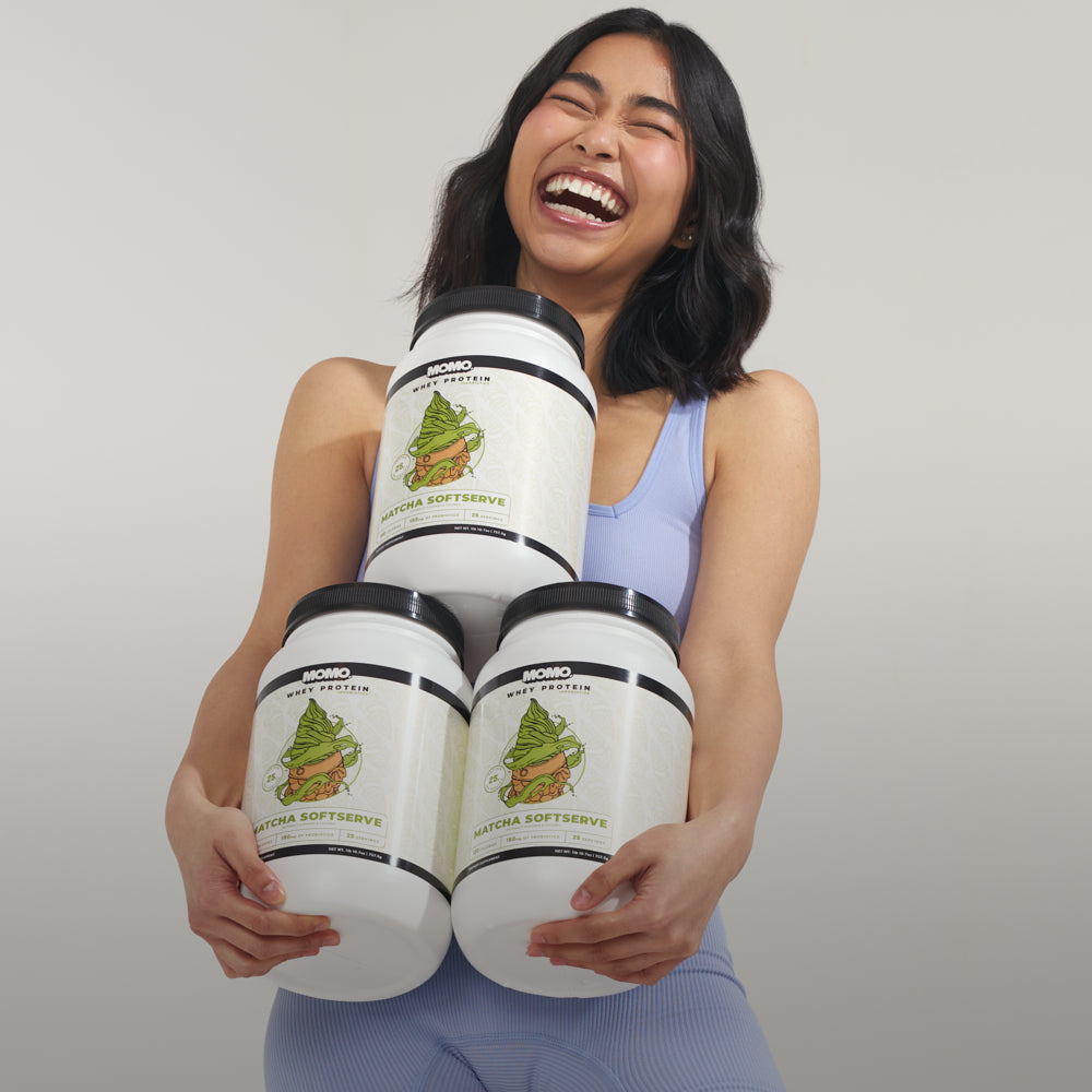 Asian girl laughing with 3 tubs of Matcha Softserve Whey Protein