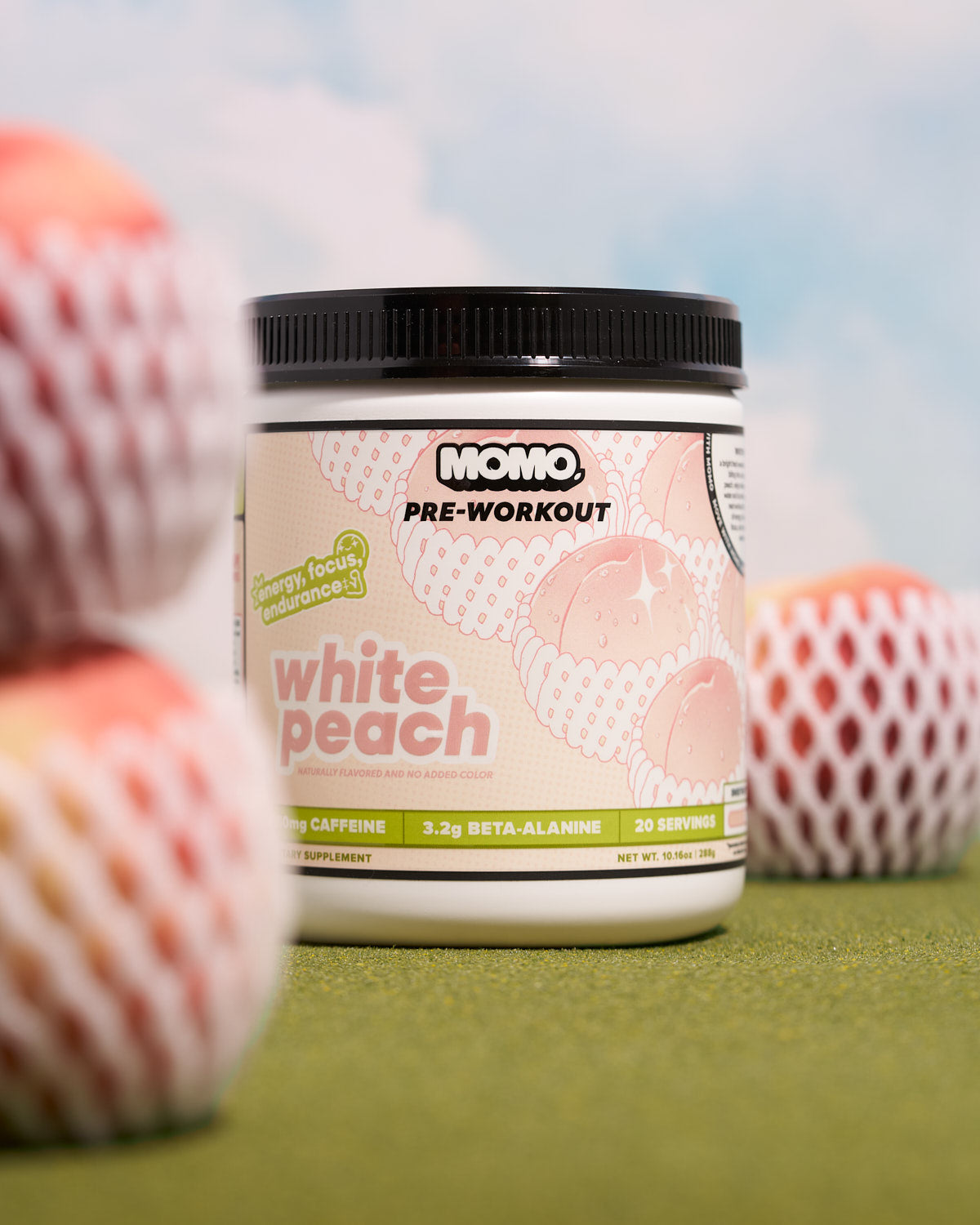 MOMO White Peach Pre-Workout on green grass and blue sky, surrounded by peaches in nets