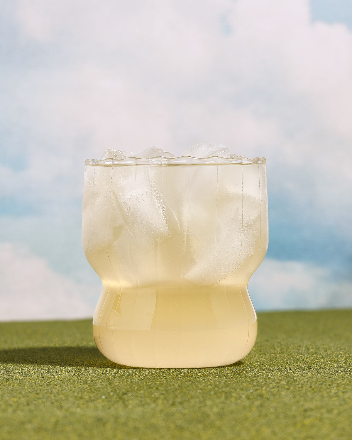 MOMO White Peach Pre-workout in a refreshing clear glass with ice. Sitting on grass with a blue sky behind.